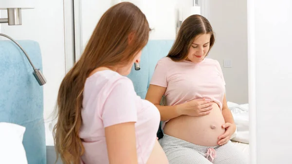 Vacker leende gravid kvinna sitter på sängen och tittar i sin spegelbild på stora spegeln. Vacker graviditet och förväntan på barnet. — Stockfoto