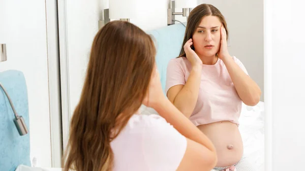 Portrét těhotné ženy trpící bolestí hlavy, jak se dívá do zrcadla. Koncepce těhotenství zdravotní a lékařské vyšetření. — Stock fotografie