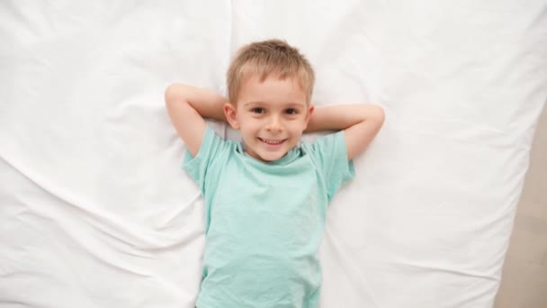 Портрет улыбающегося мальчика, лежащего на белых простынях на кровати и смотрящего в камеру. Концепция счастливых детей, хорошо проводящих время дома — стоковое видео