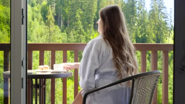 バスローブの美しいブルネットの女性は、ホテルの部屋のテラスに座って朝食を持っています。夏の山の中での観光と休暇の概念 — ストック動画