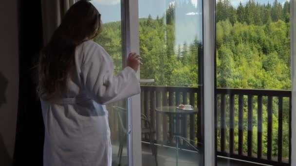 Belle touriste brune en peignoir marchant sur la terrasse de la chambre d'hôtel et regardant sur les sommets de haute montagne et la forêt de pins. Concept de tourisme et vacances en montagne en été — Video