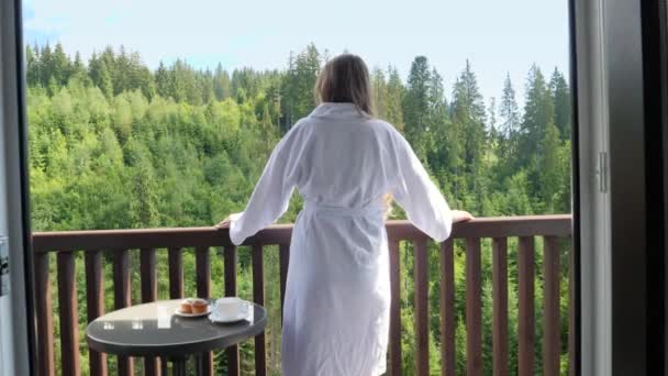 Piękna młoda kobieta w szlafroku stoi na hotelowym tarasie lub balkonie i patrzy na las sosnowy i góry. Pojęcie turystyki i wakacji w górach w lecie — Wideo stockowe