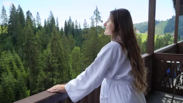 Jovem mulher relaxada de roupão de banho em pé na varanda do quarto de hotel e olhando para as montanhas e a floresta. Conceito de turismo e férias em montanhas no verão — Vídeo de Stock