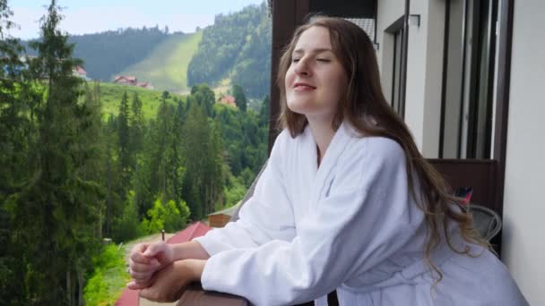 ホテルのテラスに立って、高い山を見てバスローブで幸せな笑顔の女性。夏の山の中での観光と休暇の概念 — ストック動画