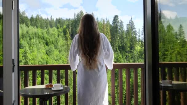Zadní pohled na šťastnou mladou ženu v županu, jak natahuje ruce poté, co se ráno probudila v horském hotelu. Koncept cestovního ruchu a dovolené v horách v létě — Stock video