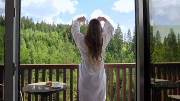 Vista posteriore di bella giovane donna che si sveglia in hotel e stende le mani sul balcone o sulla terrazza. Concetto di turismo e vacanza in montagna in estate — Video Stock