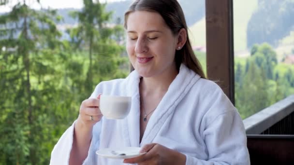 Χαρούμενο χαμόγελο woan σε μπουρνούζι πίνοντας καφέ και απολαμβάνοντας θέα στα βουνά στο ξενοδοχείο. Έννοια των ανθρώπων που ταξιδεύουν, τον τουρισμό και τις διακοπές στα βουνά το καλοκαίρι — Αρχείο Βίντεο