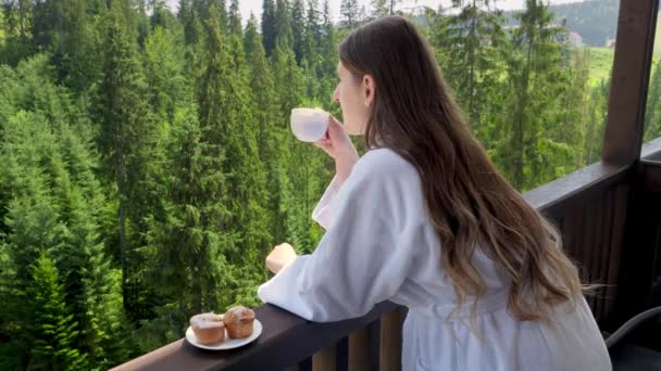 バスローブを着た若い女性が朝起きて、ホテルの部屋でバルコニーで朝食を取っています。夏の山の中で旅行、観光、休暇の人々の概念 — ストック動画