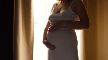 Gecelikli, güzel hamile bir kadının büyük bir pencereden parlayan parlak güneşe karşı durduğu ve karnını okşadığı yakın plan. Mutlu hamilelik ve bebek sahibi olma kavramı