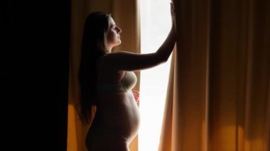 Güzel, gülümseyen hamile bir kadın perdeleri açar ve otel odasının büyük penceresinde durur. Mutlu hamilelik ve bebek beklentisi kavramı