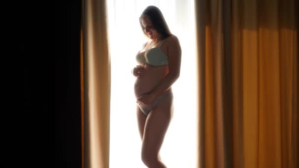 Mosolygó terhes nő fehérneműben pózol a nagy ablakban a hálószobában, és simogatja a növekvő hasát. A boldog terhesség és a babaelőrejelzés fogalma — Stock videók
