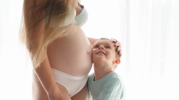 A fiatal terhes anya ölelgeti és simogatja a kisfiát, ahogy átöleli a hálószobában lévő nagy ablakban. A családi boldogság, a szeretet és a baba-várakozás fogalma — Stock videók
