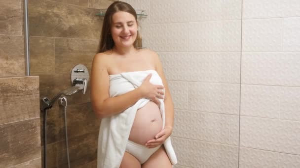 Szczęśliwa uśmiechnięta ciężarna kobieta pokryta ręcznikiem głaskająca się po brzuchu przed wzięciem prysznica w ośrodku hotelowym. Pojęcie kobiecej urody, pielęgnacji ciała i opieki zdrowotnej w czasie ciąży. — Wideo stockowe