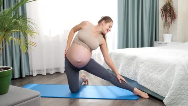 Αργή κίνηση της νεαρής χαμογελαστής εγκύου που τεντώνεται και ζεσταίνεται πριν κάνει γυμναστική στο σπίτι. Έννοια της υγειονομικής περίθαλψης και του αθλητισμού κατά τη διάρκεια της εγκυμοσύνης. — Αρχείο Βίντεο