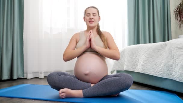 Bela mulher grávida sorridente meditando no tapete de fitness em grande janela e acariciando sua barriga grande. Conceito de estilo de vida saudável, cuidados de saúde e esportes durante a gravidez — Vídeo de Stock