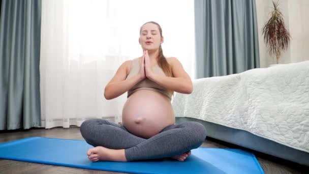 Lykkelig smilende gravid kvinde, der praktiserer yoga og mediterer strøg hendes store mave på stort vindue derhjemme. Begrebet sund livsstil, sundhedspleje og sport under graviditeten – Stock-video