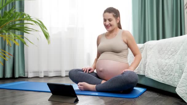 Красива вагітна жінка розтягується і виконує фізичні вправи на онлайн-урок йоги за допомогою планшетного комп'ютера. Концепція здорового способу життя, охорони здоров'я та спорту під час вагітності — стокове відео