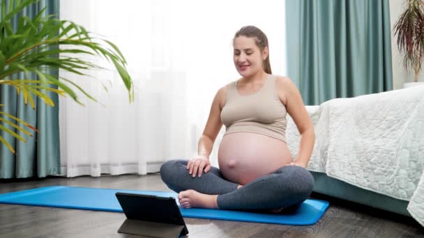 Usmívající se těhotná žena praktikující jógu online při pohledu na tablet počítače. Koncepce zdravého životního stylu, zdravotní péče a sportu během těhotenství — Stock video