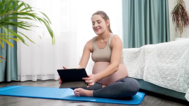 Mujer embarazada joven sentada en la esterilla de fitness y practicando yoga en línea en la tableta. Concepto de estilo de vida saludable, atención médica y deportes durante el embarazo — Vídeos de Stock