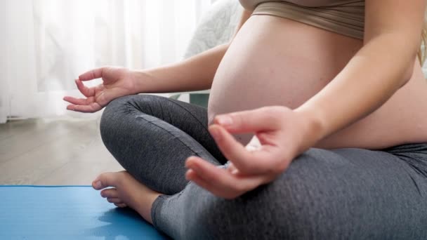 CLoseup de la mujer embarazada doblando los dedos y sentada en la postura del yoga del loto en la estera de la aptitud en ventana grande. Concepto de estilo de vida saludable, atención médica y deportes durante el embarazo — Vídeos de Stock