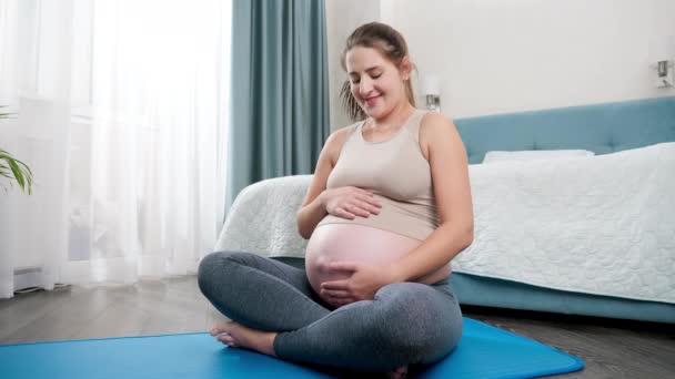 Porträtt av vacker gravid kvinna smeker hennes stora mage efter att ha tränat yoga och fitness på matta hemma. Begreppet hälsosam livsstil, hälsovård och idrott under graviditeten — Stockvideo