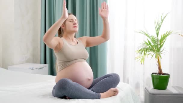 Mooie jonge zwangere vrouw die mediteert en yoga oefeningen doet terwijl ze naast het grote raam op bed zit. Concept van gezonde levensstijl, gezondheidszorg en sport tijdens de zwangerschap — Stockvideo