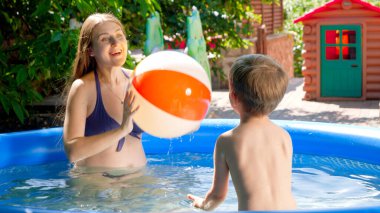 Mutlu gülümseyen anne ve küçük çocuk evin arka bahçesinde şişme yüzme havuzunda plaj topuyla oynuyor. Aile yaz tatili ve tatiller