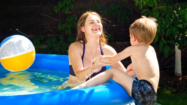 Pequeño niño sonriente escalando en la piscina inflable en el patio trasero de la casa y jugando con la madre joven. Vacaciones familiares de verano y vacaciones — Foto de Stock