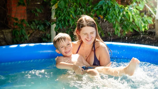 Feliz madre sonriente sosteniendo y apoyando a su pequeño hijo aprendiendo a nadar en la piscina inflable. Vacaciones familiares de verano y vacaciones — Foto de Stock