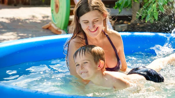 Joven madre sonriente sosteniendo y apoyando su pequeña piscina en el soleado día de verano. Familia con niños vacaciones de verano y vacaciones. — Foto de Stock