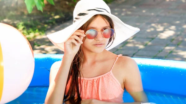 漂亮的少女戴着夏帽，戴着时髦的太阳镜，在自家后院的充气游泳池里悠闲自在地笑着。快乐暑假及假期的概念 — 图库照片