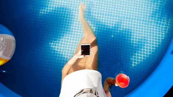 Top uitzicht op mooie slanke vrouw met cocktail zwemmen en ontspannen in het buitenzwembad en met behulp van smartphone tijdens het surfen op internet of sociale media. Concept van gelukkige zomervakantie en vakantie — Stockfoto