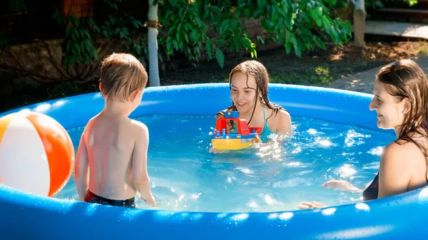 Uśmiechnięty chłopczyk bawiący się z matką isster ze statkiem-zabawką w nadmuchiwanym basenie w ogrodzie podwórkowym. Pojęcie szczęśliwej i wesołej rodziny letnie wakacje i wakacje — Zdjęcie stockowe