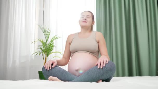 Vrolijk lachende zwangere vrouw die yoga en meditatie beoefent tegen groot raam en heldere ochtendzon. Concept van gezonde levensstijl, gezondheidszorg en sport tijdens de zwangerschap — Stockvideo