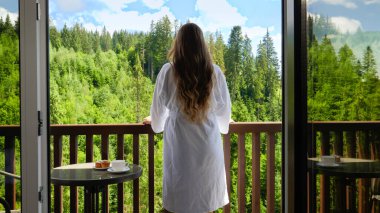 Güzel genç bir kadın sabah uyanıyor ve ormanı ve dağları izlerken otel odasına ellerini uzatıyor. Turizm kavramı ve yazın dağlarda tatil