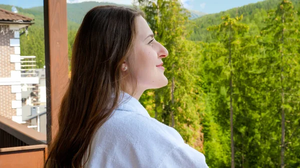 ホテルの部屋から山や森の景色を楽しむバスローブの美しい笑顔の女性の肖像画。夏の山の中での観光と休暇の概念 — ストック写真