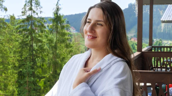 Heureuse femme souriante en peignoir debout sur la terrasse de l'hôtel et regardant les hautes montagnes. Concept de tourisme et vacances en montagne en été — Photo