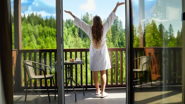 Rear view of happy young woman in bathrobe stretching out hands after waking up in morning at mountain hotel. Concept of tourism and vacation in mountains at summer — Stock Photo, Image