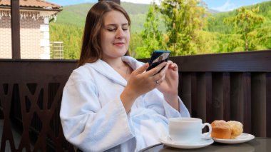 Güzel, gülümseyen bir kadın kahvaltı yapıyor ve otel odasının balkonunda akıllı telefon kullanıyor. Yaz aylarında dağlarda seyahat, turizm ve tatil kavramı