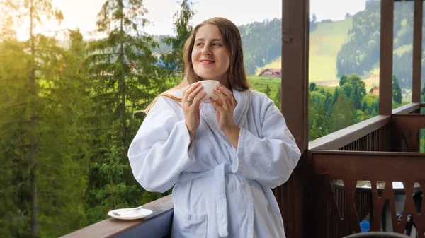 Portrait d'une jeune femme souriante en peignoir prenant le petit déjeuner et buvant du café sur le balcon ou la terrasse de la chambre d'hôtel. Concept de personnes voyageant, tourisme et vacances en montagne en été — Photo