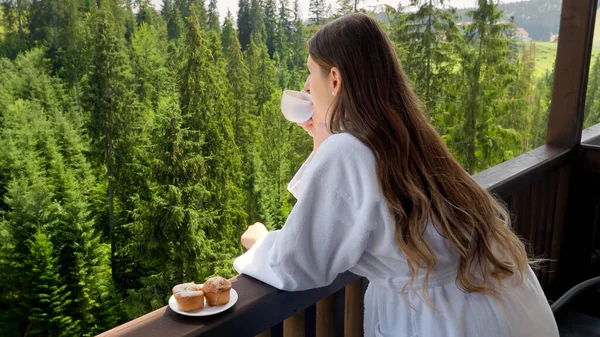 Mujer joven en albornoz despertando por la mañana y desayunando en el balcón en la habitación del hotel. Concepto de personas viajando, turismo y vacaciones en las montañas en verano —  Fotos de Stock
