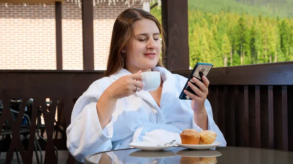 Jonge mooie vrouw in badjas ontbijten in de ochtend en met behulp van smartphone. — Stockfoto