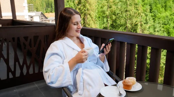 Young smiling woman in bathrobe sitting on hotel balcony and using smartphone while having breakfast. Concept of people traveling, tourism and vacation in mountains at summer — Stock Photo, Image