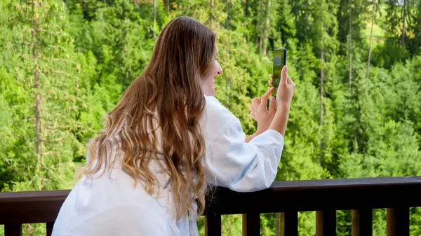 Hermosa joven en albornoz de pie en el balcón y hacer imagen de las montañas y el bosque en su teléfono inteligente. —  Fotos de Stock