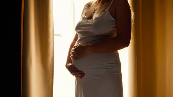 Primer plano de la hermosa mujer embarazada en vestido de noche de pie contra el sol brillante que brilla a través de una gran ventana y acariciando su gran vientre. Concepto de embarazo feliz y esperar un bebé — Foto de Stock