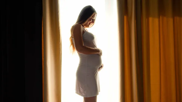 Krásná usměvavá těhotná žena stojí u velkého okna a hladí ji po břiše. Koncept šťastného těhotenství a očekávání dítěte — Stock fotografie