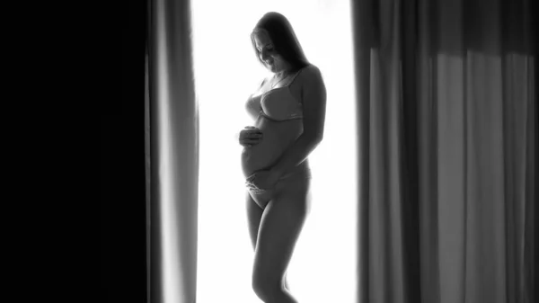 Czarno-biały obraz uśmiechniętej ciężarnej kobiety w bieliźnie, pozującej w dużym oknie w sypialni i głaskającej jej rosnący brzuch. Pojęcie szczęśliwej ciąży i antycypowania dziecka — Zdjęcie stockowe