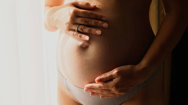 Hamile bir kadının yatak odasındaki büyük pencerenin yanında karnına nazikçe dokunup okşaması. Mutlu hamilelik ve bebek beklentisi kavramı — Stok fotoğraf