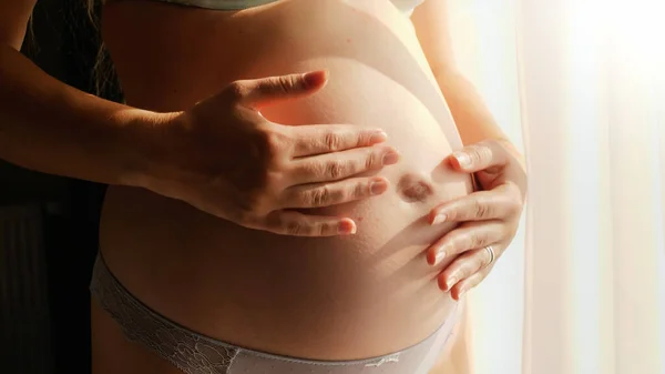 Primer plano de la mujer embarazada de pie en la luz del sol y suavemente acariciando su gran vientre. Concepto de embarazo feliz y anticipación del bebé —  Fotos de Stock