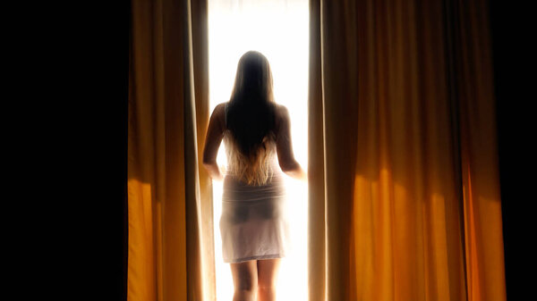 Beautiful brunette woman in night gown waking up at morning and opening curtains covering big window at hotel